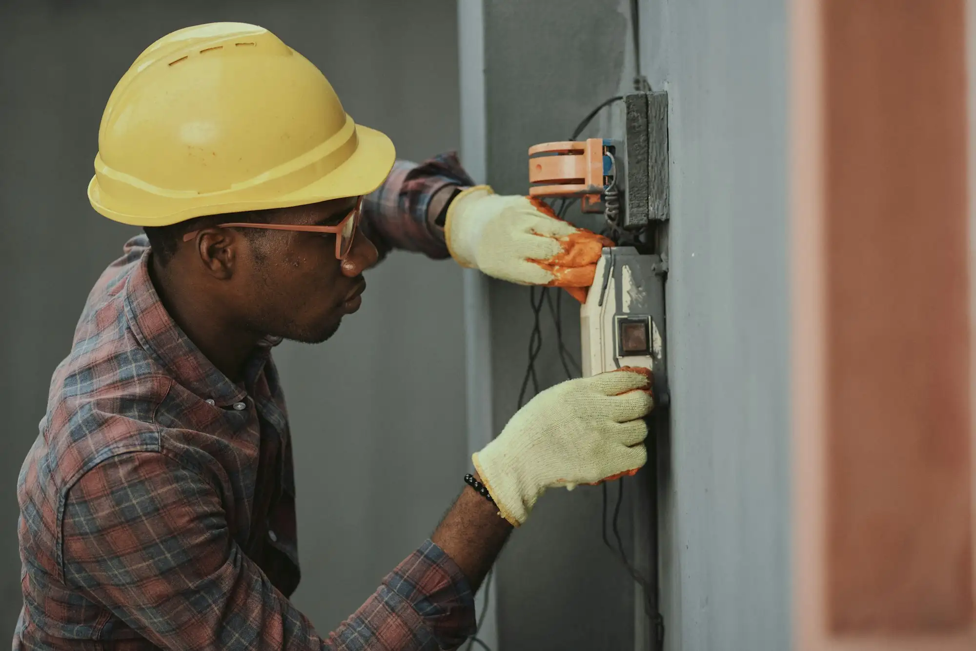 Electrical subcontractor in a yellow hard hat and safety glasses wiring a meter box on a building site