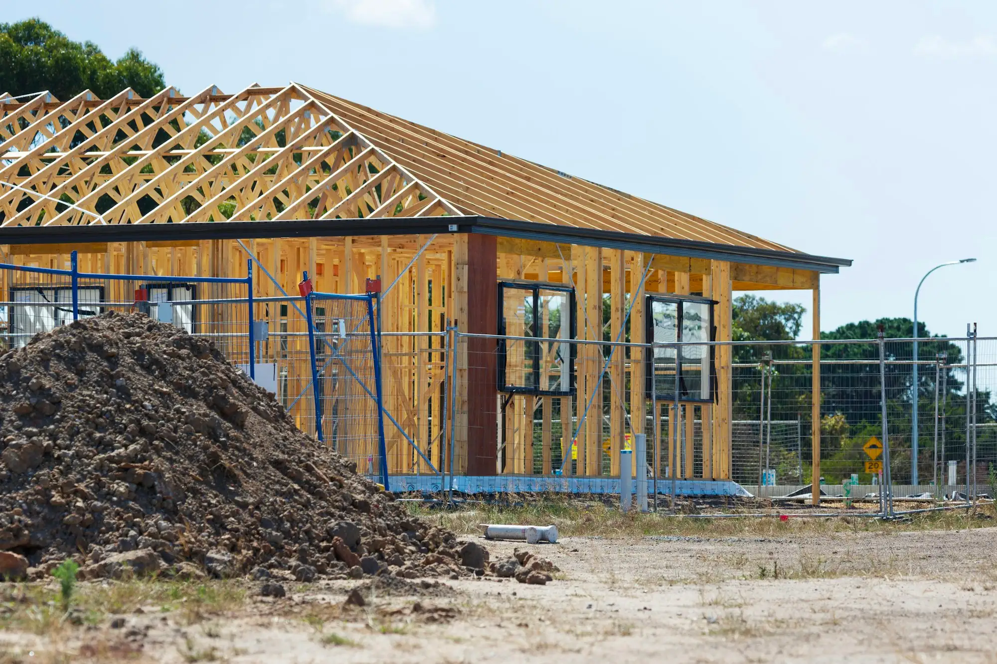 Single storey timber house frame with roof trusses behind temporary site fencing on a new Australian housing estate